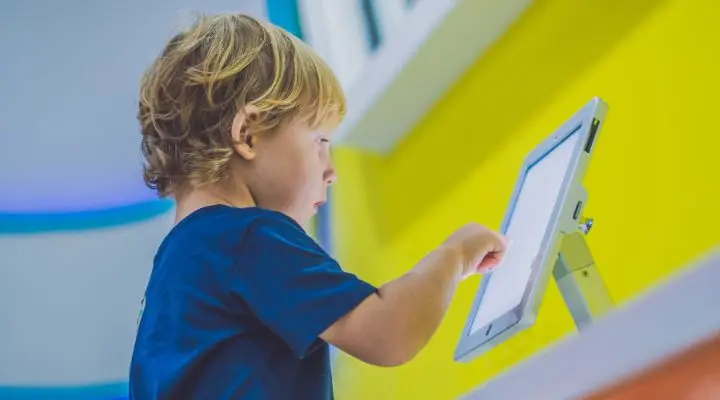 Un niño de cabello rubio, parte de la Generación Beta, interactúa con una tableta montada en un soporte, sobre un fondo amarillo y blanco vibrante.