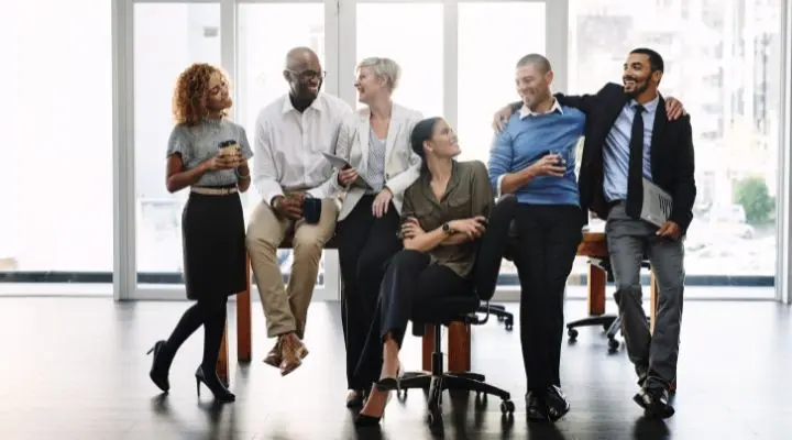 Un grupo de seis personas vestidas de negocios se reúnen en una oficina, algunas sentadas en escritorios, conversando y sosteniendo tazas de café, centrándose en estrategias para la retención del talento humano.