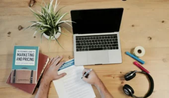 Una persona escribe en un documento en un escritorio de madera con una computadora portátil, libros, una planta, auriculares, un teléfono inteligente, bolígrafos y cinta adhesiva.