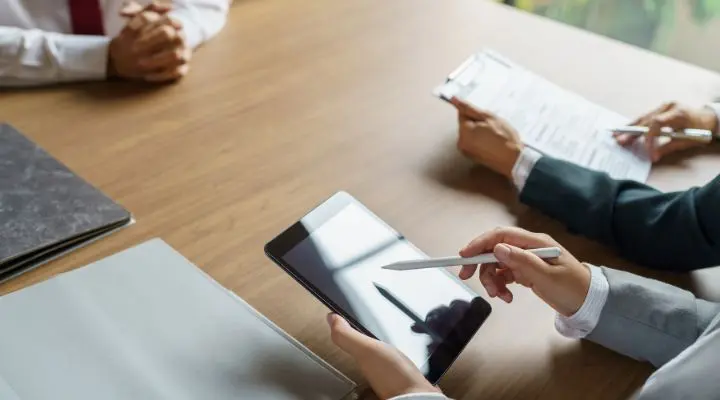 En una mesa, tres profesionales vestidos de negocios discuten sobre vacantes laborales; uno sostiene una tableta, otro revisa papeles, mientras que el tercero está sentado con las manos entrelazadas, sumido en sus pensamientos.