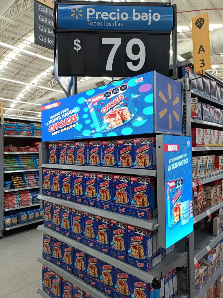 Un vibrante escaparate de supermercado muestra cajas de cereales con un precio de $79, realzadas con carteles promocionales en español. Con Walmart Connect ampliando el alcance, los estantes están prolijamente abastecidos bajo un acogedor cartel de "Precio bajo".