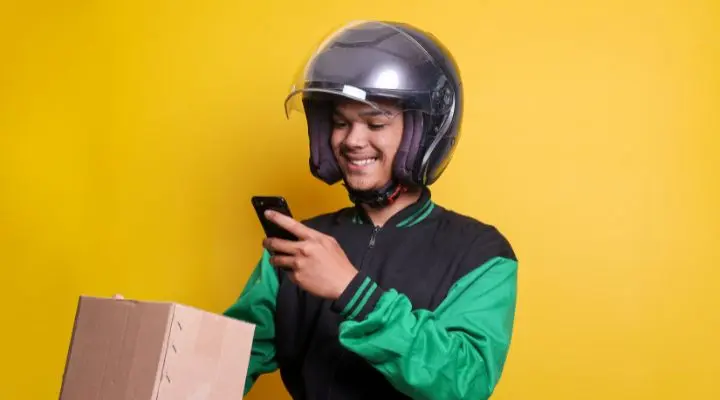 Una persona con casco y chaqueta sostiene un teléfono inteligente y una caja de cartón, encarnando la eficiencia de las plataformas digitales sobre un fondo amarillo vibrante.