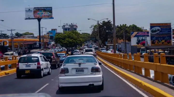 Los coches se agolpan en medio del tráfico en una calle de la ciudad, rodeados de barreras de color amarillo brillante y edificios imponentes. Los carteles publicitarios se ciernen sobre sus cabezas mientras los árboles bordean la carretera, creando un vibrante cuadro urbano.