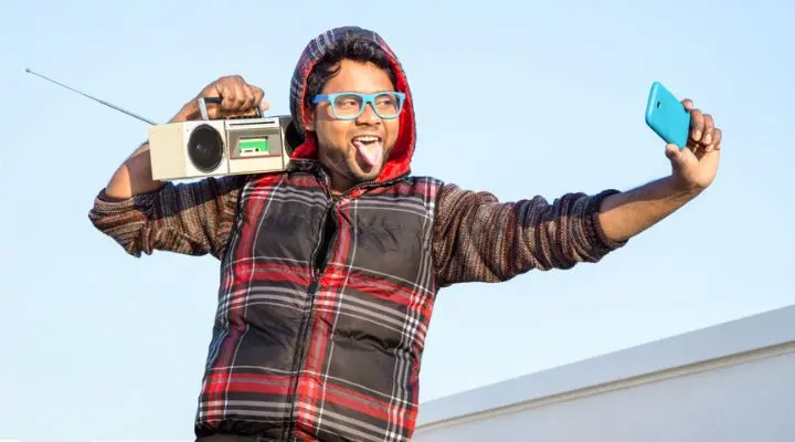 Una persona con gafas y un chaleco a cuadros posa con una radio, un estéreo portátil en el hombro y un teléfono inteligente para una selfie.