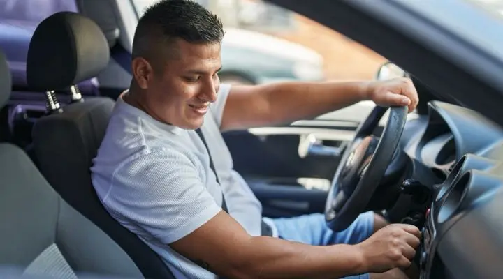 Un hombre con camisa blanca y pantalones cortos está sentado en un automóvil, sonriendo mientras gira la llave de encendido, ansioso por que sus canciones favoritas suenen en la radio.