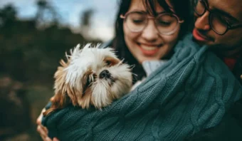 Una pareja sostiene un perrito peludo envuelto en una manta en el exterior. Ambos sonríen y llevan gafas.