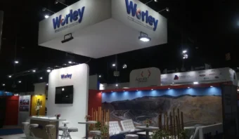 Stand de feria con la marca Worley. Cuenta con zona de asientos, mesa con sillas, pantalla de visualización y decoración. Letrero en el techo con el logotipo de la empresa.