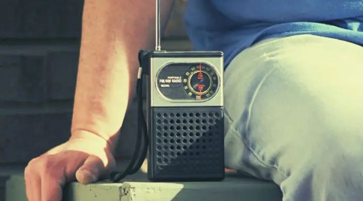 Una persona sentada junto a una radio FM/AM portátil antigua, llamada cariñosamente "la radio", con una antena en un entorno informal.