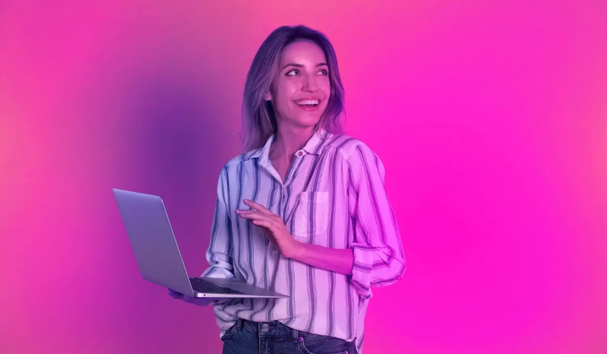 Una persona con una camisa a rayas sostiene una computadora portátil y sonríe contra un fondo degradado de color rosa y morado.