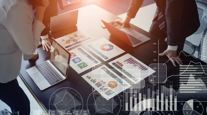 Personas alrededor de una mesa con computadoras portátiles y gráficos coloridos, discutiendo análisis de datos y planificación de medios. Gráficos digitales superpuestos a la imagen indican análisis financieros y estrategia empresarial.