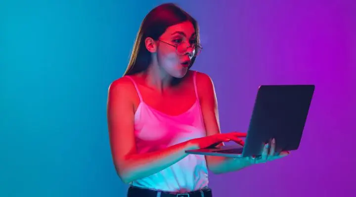 Las mujeres amantes de la tecnología apreciarán la escena: una mujer con gafas parece sorprendida mientras escribe en su portátil, sobre un fondo azul y morado vibrante.