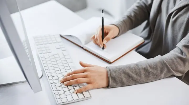 Una persona anota mensajes clave en un cuaderno con una mano, mientras la otra descansa ligeramente sobre el teclado de una computadora.