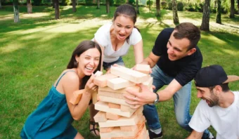 Cuatro personas al aire libre jugando con grandes bloques de madera sobre el césped, pareciendo emocionadas y comprometidas.
