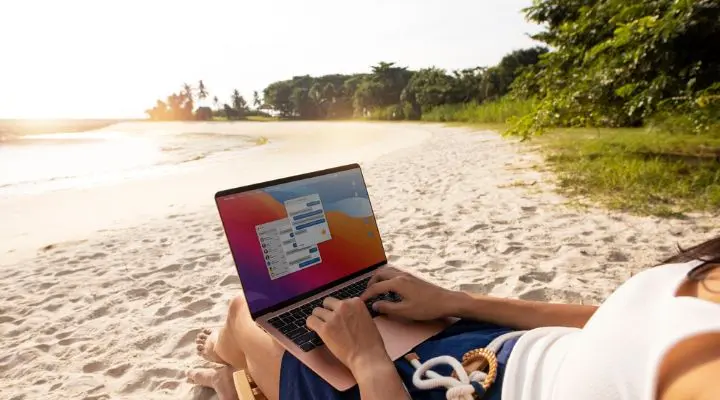 En una playa de arena, una persona teclea en una laptop, creando campañas digitales mientras el sol se pone sobre el océano. Los frondosos árboles verdes se mecen suavemente al fondo, realzando este idílico entorno de teletrabajo.