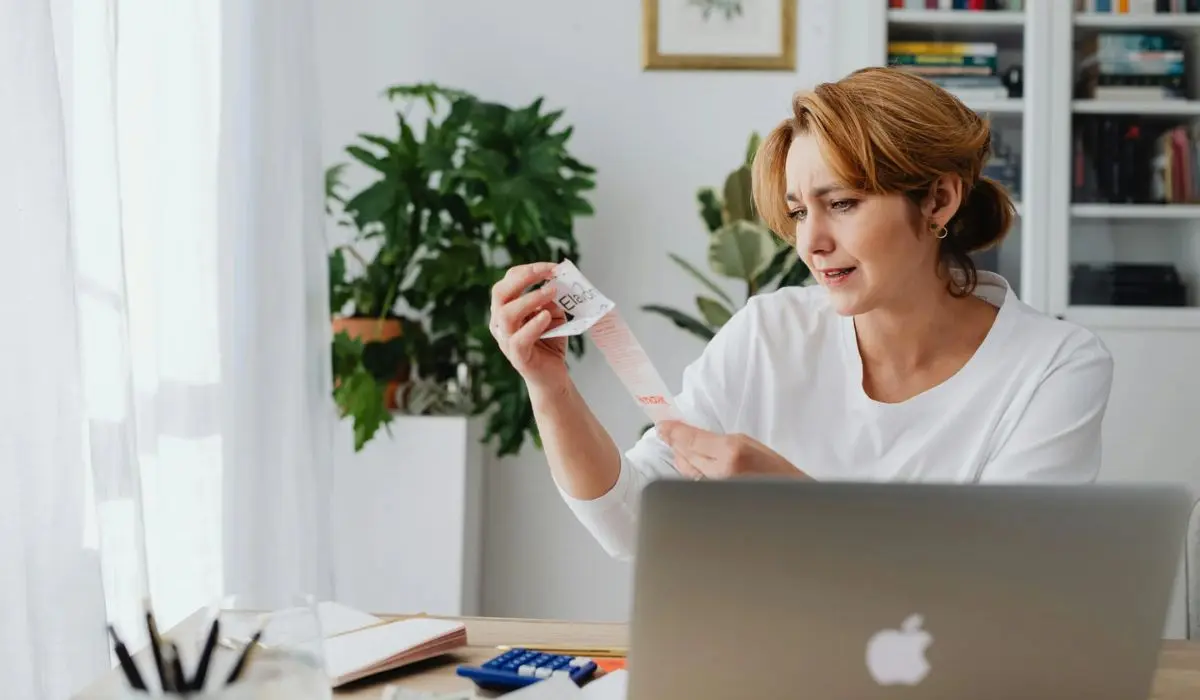 Una mujer está sentada en un escritorio con una laptop, sosteniendo y leyendo un recibo. Al fondo hay plantas, libros y material de oficina.