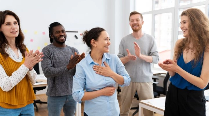 Cinco personas en una sala iluminada sonríen y aplauden, celebrando un momento de liderazgo. La mujer del centro, personificando el verdadero liderazgo, luce conmovida al llevarse la mano al pecho.