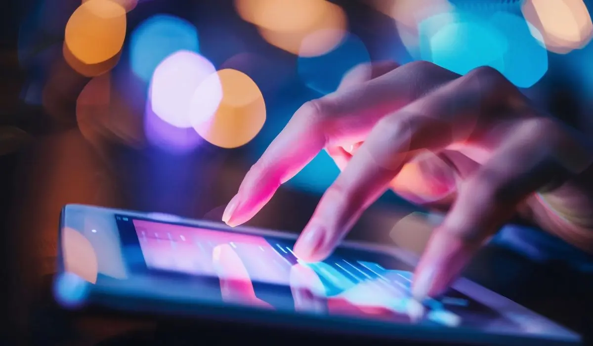 Un primer plano de una mano que utiliza una tableta con pantalla táctil, con luces borrosas de colores en el fondo.