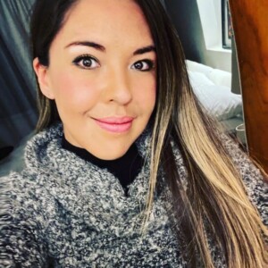 Una mujer con cabello largo y liso y un suéter gris se toma una selfie en un interior, con una cama y una ventana al fondo.