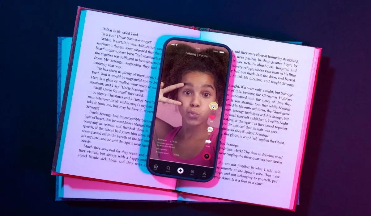 Teléfono inteligente que muestra a una persona haciendo el signo de la paz en una aplicación de video, apoyada sobre un libro abierto con iluminación de colores.