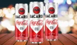 Tres latas de Bacardi mezcladas con Coca-Cola, cada una de 355 ml con 5% de alcohol por volumen, expuestas sobre una superficie de madera con un fondo colorido borroso.
