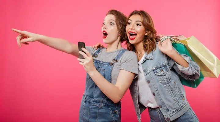 Dos mujeres miran emocionadas en la misma dirección, una sosteniendo un teléfono y la otra llevando bolsas de compras, encarnando el marketing consciente sobre un fondo rosa.