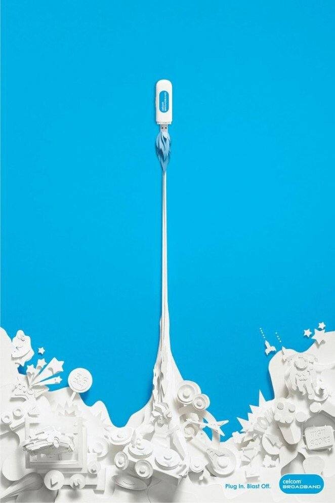 Un módem USB se representa como un cohete que se lanza hacia el cielo, dejando una estela blanca desde una ciudad de papel detallada debajo, sobre un fondo azul: un ejemplo de anuncios creativos que captan la atención.