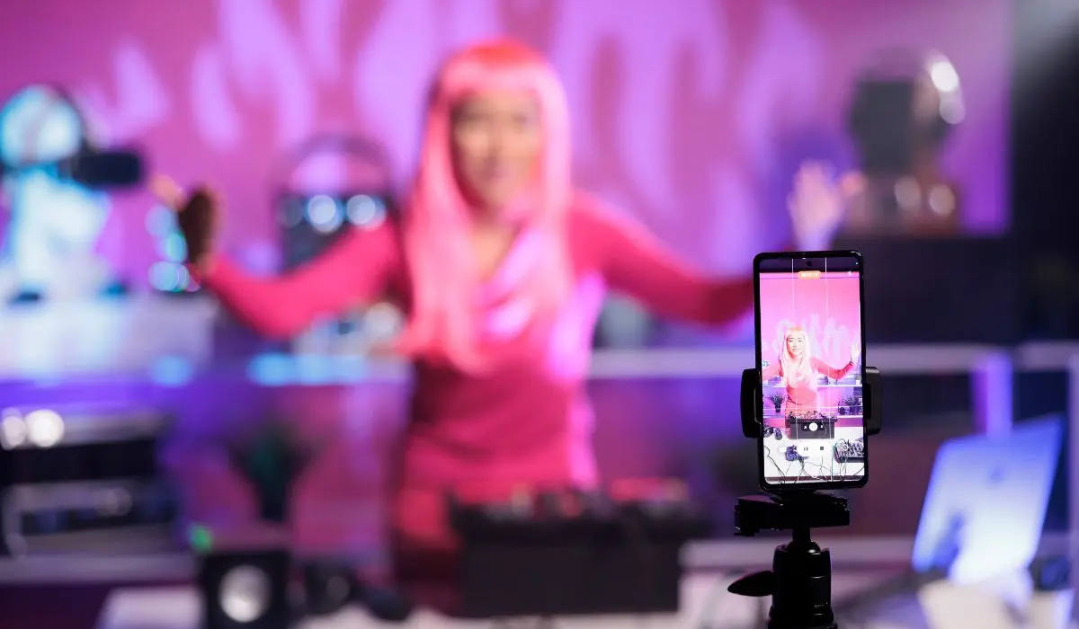 Una persona con cabello rosa y ropa rosa se encuentra detrás de un equipo de DJ, siendo grabada con un teléfono inteligente montado en un trípode en un estudio con iluminación violeta.
