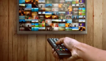 Una mano que sostiene un control remoto apunta a una pantalla de televisión que muestra una cuadrícula de varias imágenes coloridas y miniaturas de videos.