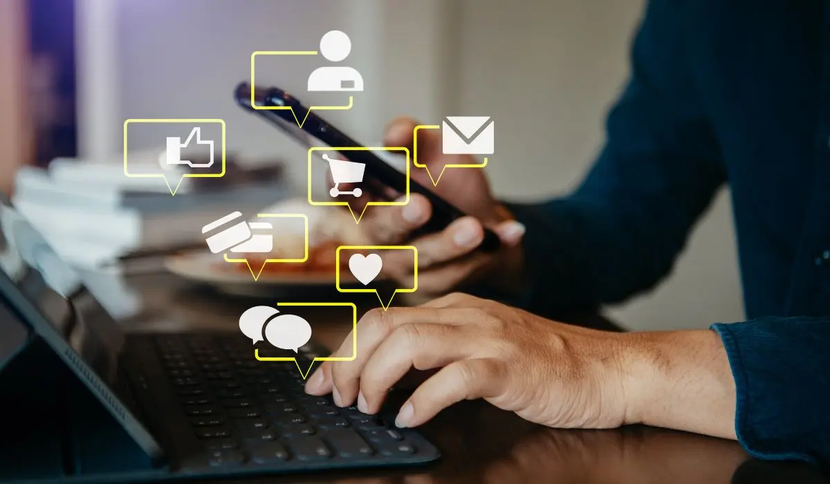 Una persona escribe en un teclado con una mano y sostiene un teléfono inteligente en la otra, rodeada de íconos ilustrados de redes sociales y comunicación.