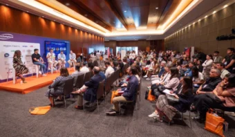 Un panel de oradores se dirige a la audiencia en el Marketplace Summit 2025, mientras los asistentes escuchan atentamente y algunos sostienen bolsas de color naranja en la sala de conferencias.