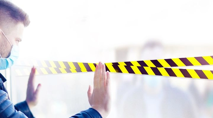 Una persona que lleva una mascarilla toca un vidrio marcado con cinta de precaución negra y amarilla, lo que sugiere negocios restringidos, mientras una figura borrosa está parada al otro lado.