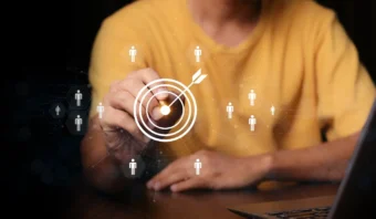 Persona con camisa amarilla tocando una diana virtual con un dardo, rodeada de iconos humanos, simbolizando la segmentación de audiencias o el marketing digital y destacando la relevancia de marca en estrategias efectivas.