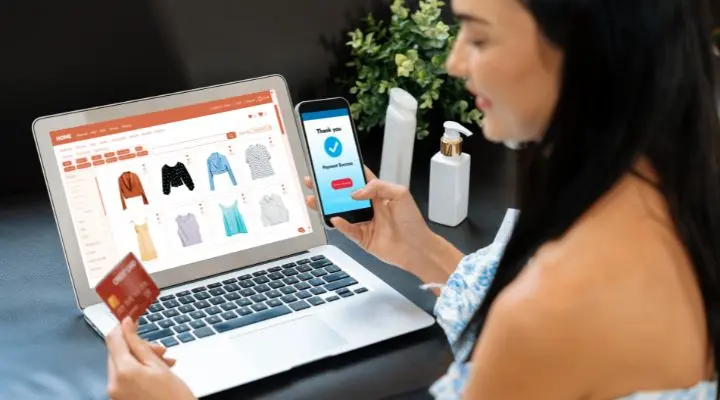 Una mujer sostiene una tarjeta de crédito y un teléfono inteligente mientras compra ropa en línea en una computadora portátil, con un mensaje de pago exitoso en la pantalla de su teléfono, mostrando que es seguro comprar por internet.