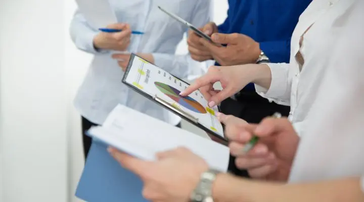 Cuatro personas sosteniendo documentos y una tableta, revisando un gráfico circular basado en encuestas públicas y tomando notas durante una reunión o discusión.