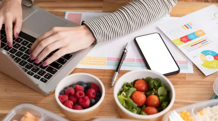 Persona escribiendo en una computadora portátil en un escritorio rodeado de un teléfono inteligente, un bolígrafo, gráficos impresos y tazones de fruta y ensalada, planificando estrategias de marketing gastronómico.