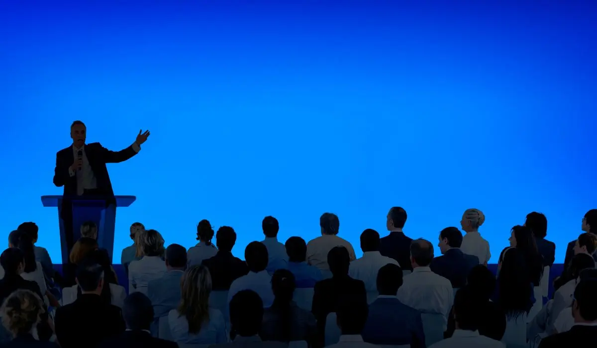 Un orador, después de tomar tiempo para preparar una conferencia, se sitúa en un podio y se dirige a una audiencia sentada en filas contra un fondo azul liso.