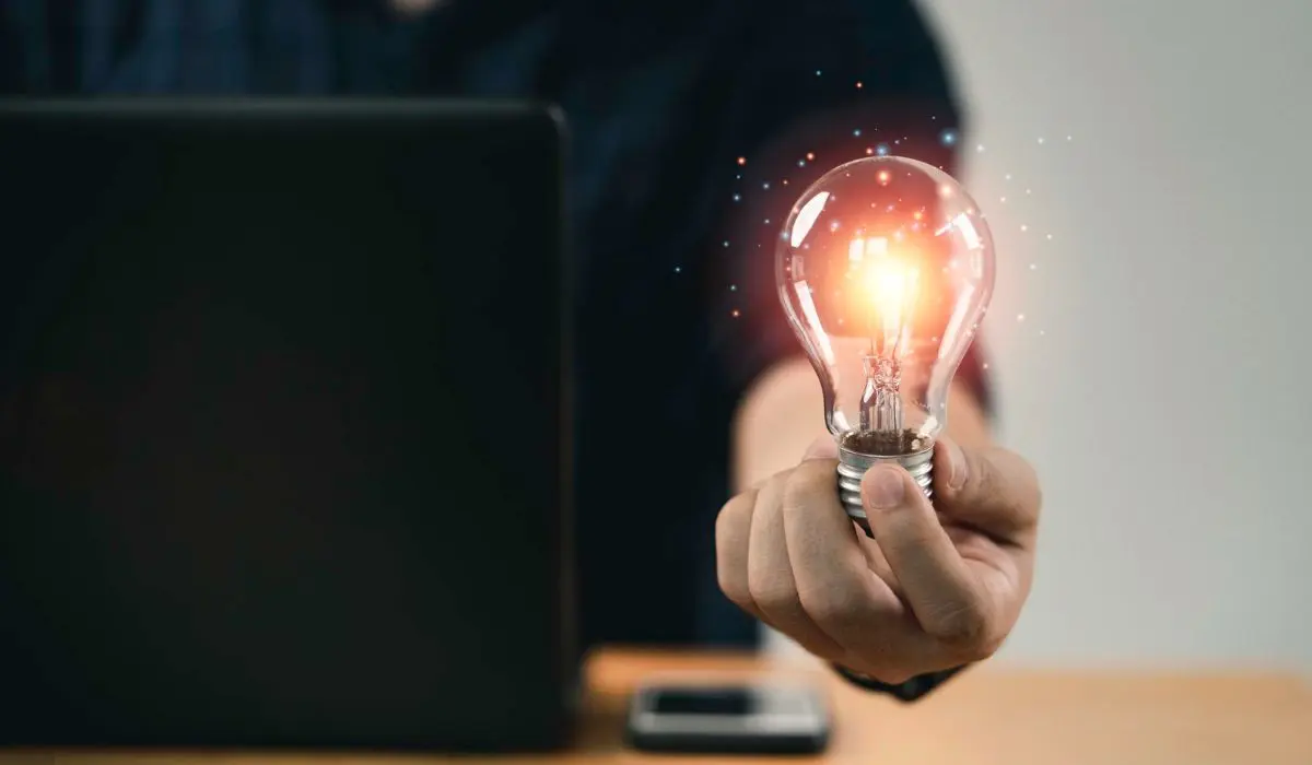 Una persona sostiene una bombilla encendida frente a una computadora portátil en un escritorio, con un teléfono inteligente al lado: listo para innovar y generar nuevas ideas.