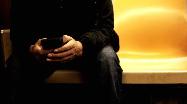 Persona sentada en un asiento amarillo del metro, sosteniendo un teléfono inteligente con ambas manos, posiblemente manejando las redes sociales de un fallecido; la parte superior del cuerpo y la cara no son visibles.
