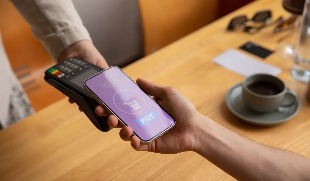 Una persona sostiene un teléfono inteligente con una aplicación de pago cerca de un lector de tarjetas para realizar un pago sin contacto; se ganan puntos de fidelidad mientras disfrutan de una taza de café y papeles en la mesa.