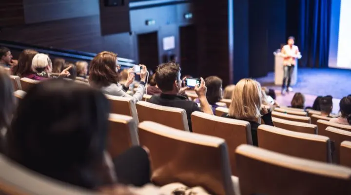 Público sentado en una sala de conferencias, algunos sosteniendo teléfonos, mientras un ponente de éxito se encuentra de pie en un podio en el escenario frente a una pantalla grande.