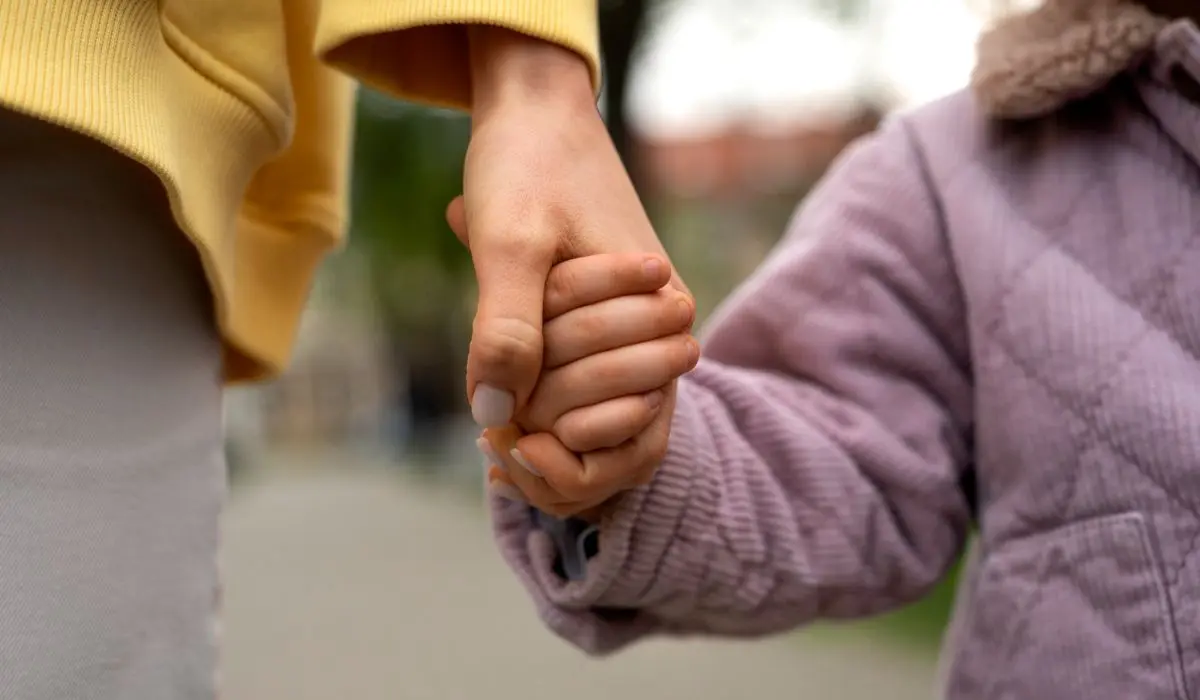 Primer plano de un adulto y un niño tomados de la mano al aire libre, simbolizando la conexión entre generaciones. El adulto lleva una manga amarilla; el niño, con una chaqueta acolchada lavanda, representa la nueva generación. El fondo está desenfocado.