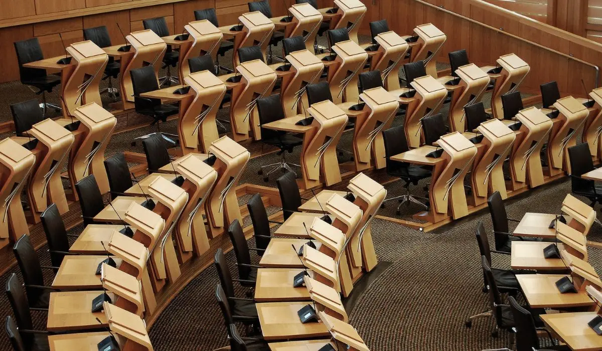 Filas de escritorios de madera vacíos y sillas negras dispuestas en un patrón curvo dentro de una moderna cámara legislativa o de conferencias, lo que refleja el potencial de reinvención política dentro de su cuidadoso diseño.