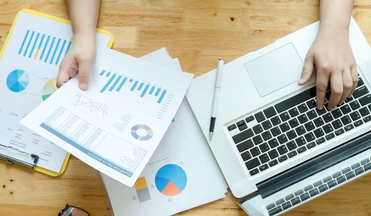 Persona que trabaja en un escritorio con una computadora portátil, sosteniendo gráficos y cuadros impresos, junto con un informe de redes sociales y documentos financieros, con un bolígrafo sobre la superficie de madera.