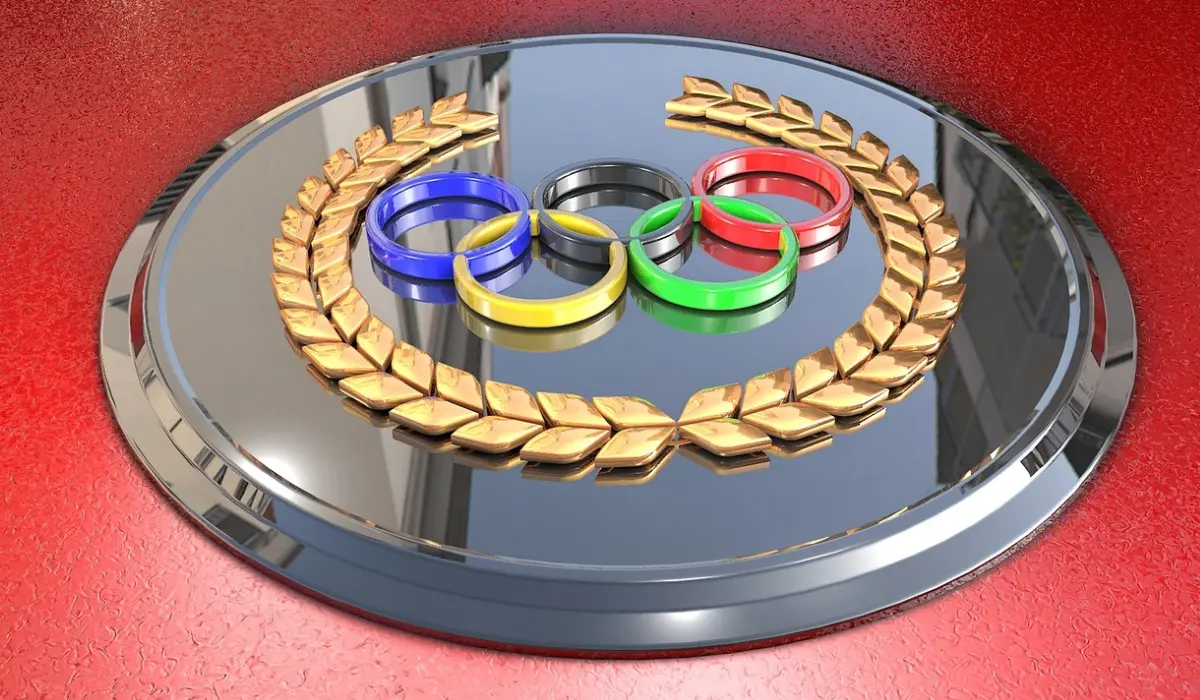 Una placa metálica que presenta los anillos olímpicos en azul, amarillo, negro, verde y rojo, que simbolizan a México en los Juegos Olímpicos, rodeada por una corona de laurel dorada y sobre una superficie texturizada de color rojo.