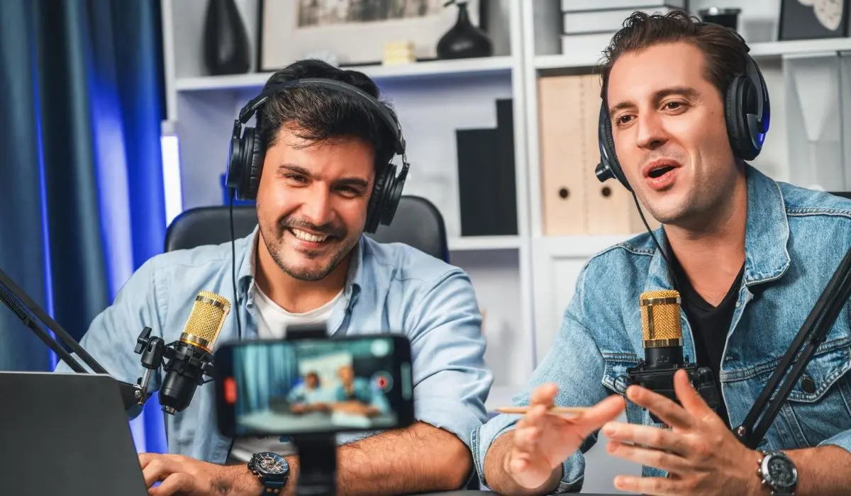 Dos hombres con auriculares hablan por micrófonos y están sentados en un escritorio con una computadora portátil, grabando un podcast o una transmisión en vivo en un estudio moderno, discutiendo el negocio de los YouTubers.