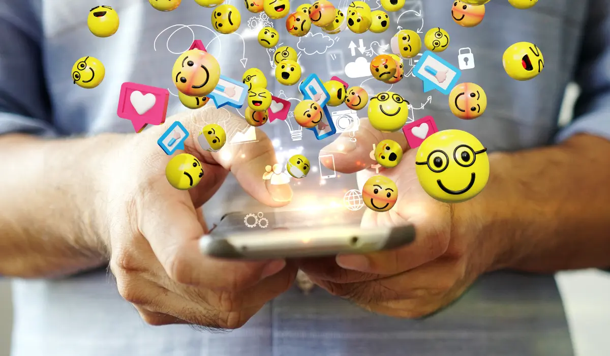 Una persona usa un teléfono inteligente mientras un emoji y varios íconos de redes sociales parecen flotar en 3D sobre el dispositivo.