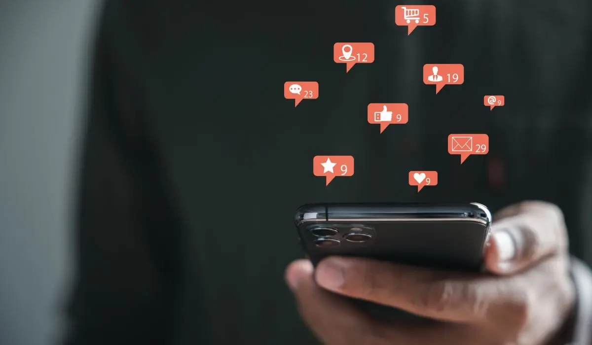 Una persona que sostiene un teléfono inteligente con múltiples íconos de notificaciones de redes sociales, incluidos Me gusta, comentarios, mensajes y solicitudes de amistad, que aparecen sobre el dispositivo, lo que resalta el alcance de las interacciones en línea.
