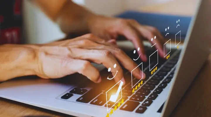 Manos escribiendo en un teclado de computadora portátil con íconos de caras sonrientes digitales y estrellas de calificación superpuestas, sugiriendo comentarios en línea o calificación de futuros clientes.