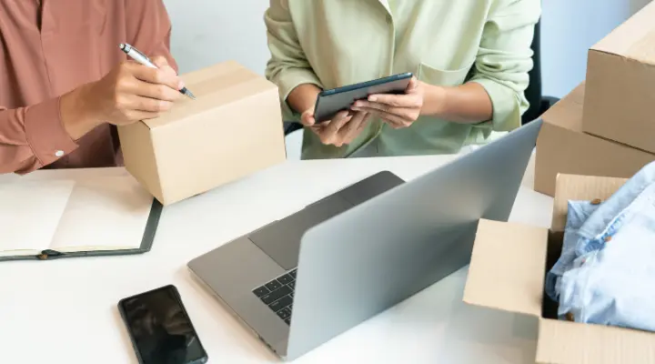 Dos personas se sientan en un escritorio con una computadora portátil, un teléfono inteligente, cuadernos, cajas de cartón y una tableta, abordando los retos de las PyMEs mientras preparan paquetes para enviar o administran un negocio en línea.