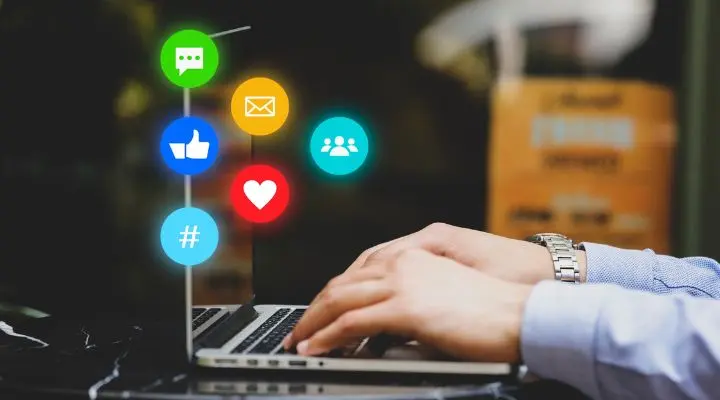 Manos de Community Management escribiendo en un teclado de computadora portátil con íconos coloridos de redes sociales, incluidos chat, me gusta, correo electrónico, hashtag y grupo, flotando sobre la pantalla: perfecto para ilustrar la gestión comunitaria en acción.